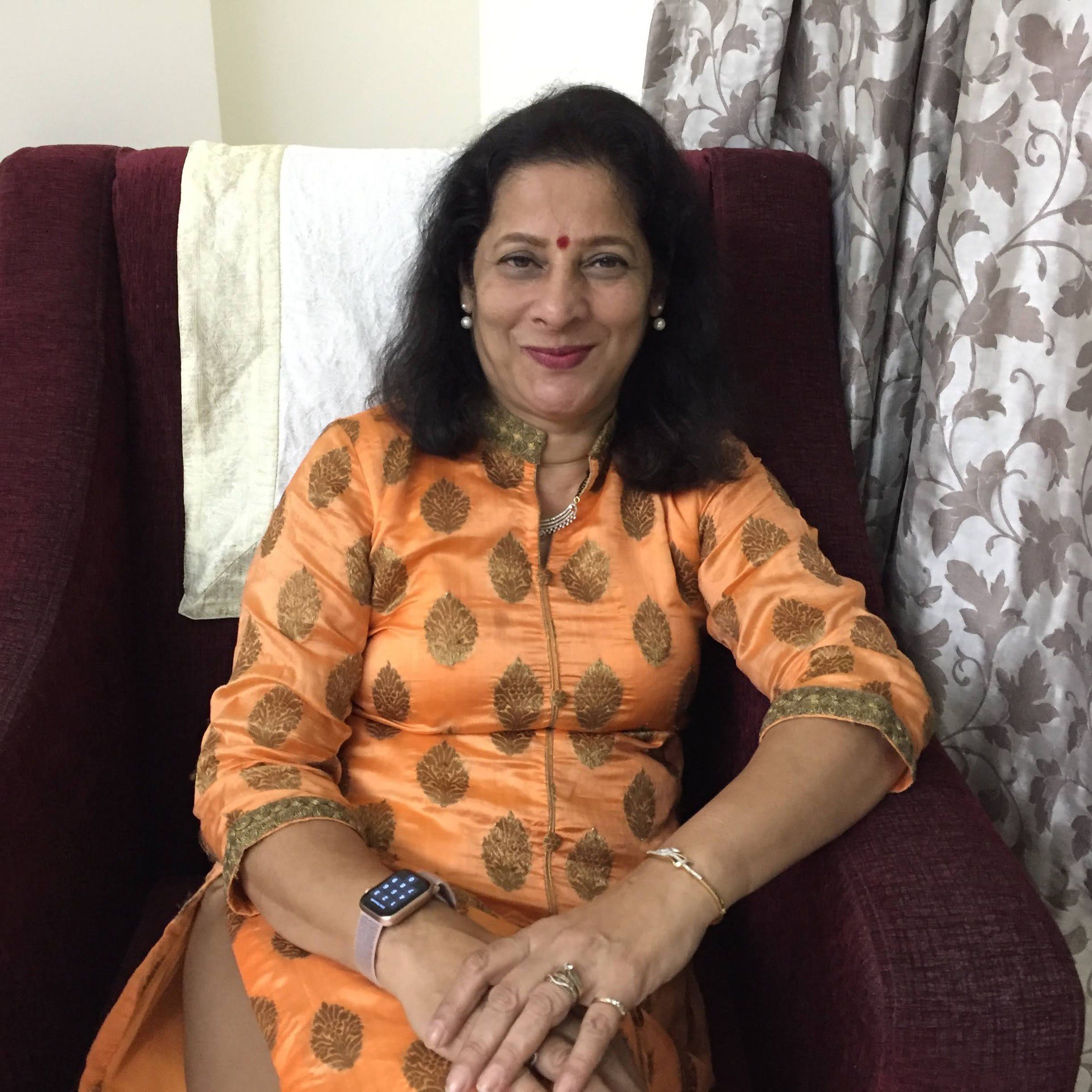 Mugdha seated in a maroon armchair at home, smiling, in an orange kurta with a gold leaf pattern