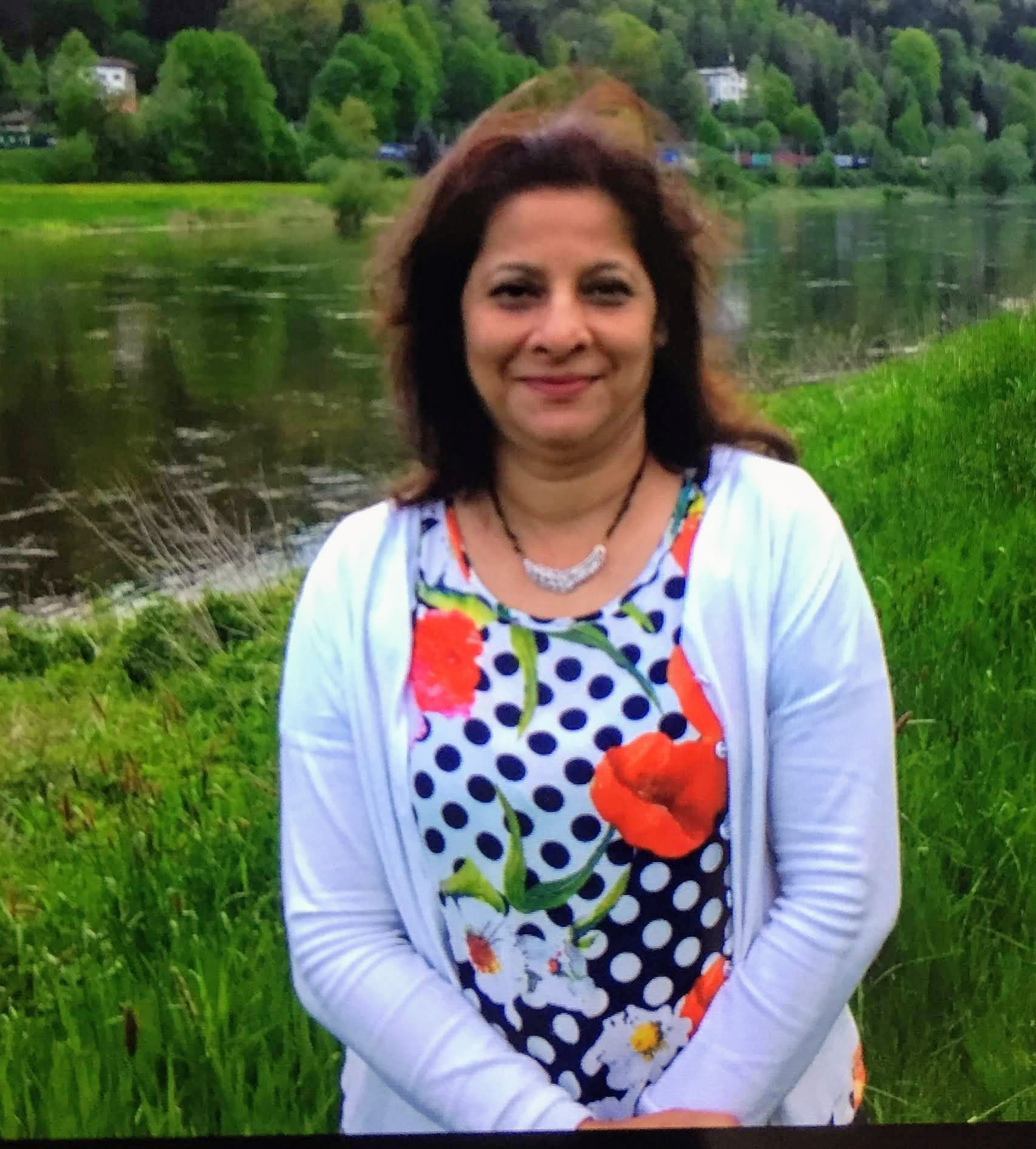 Mugdha on a grassy riverbank with a wooded hillside behind her, in a white cardigan over a polka-dot top printed with red poppies
