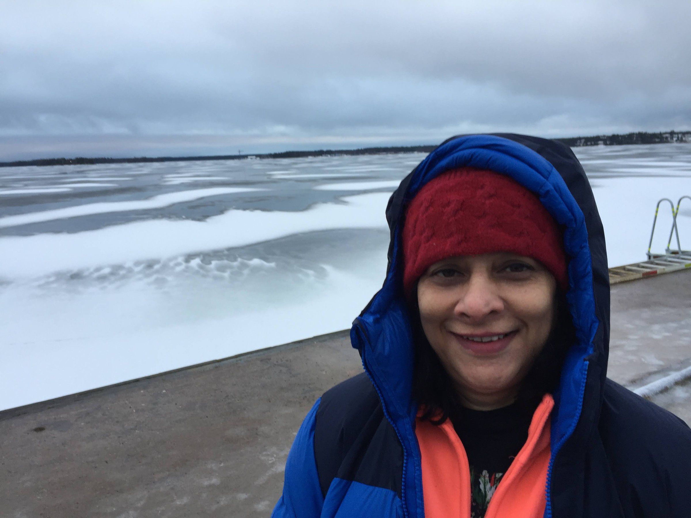 Mugdha in a blue hooded jacket and red knitted hat beside a frozen, snow-covered sea under a grey sky