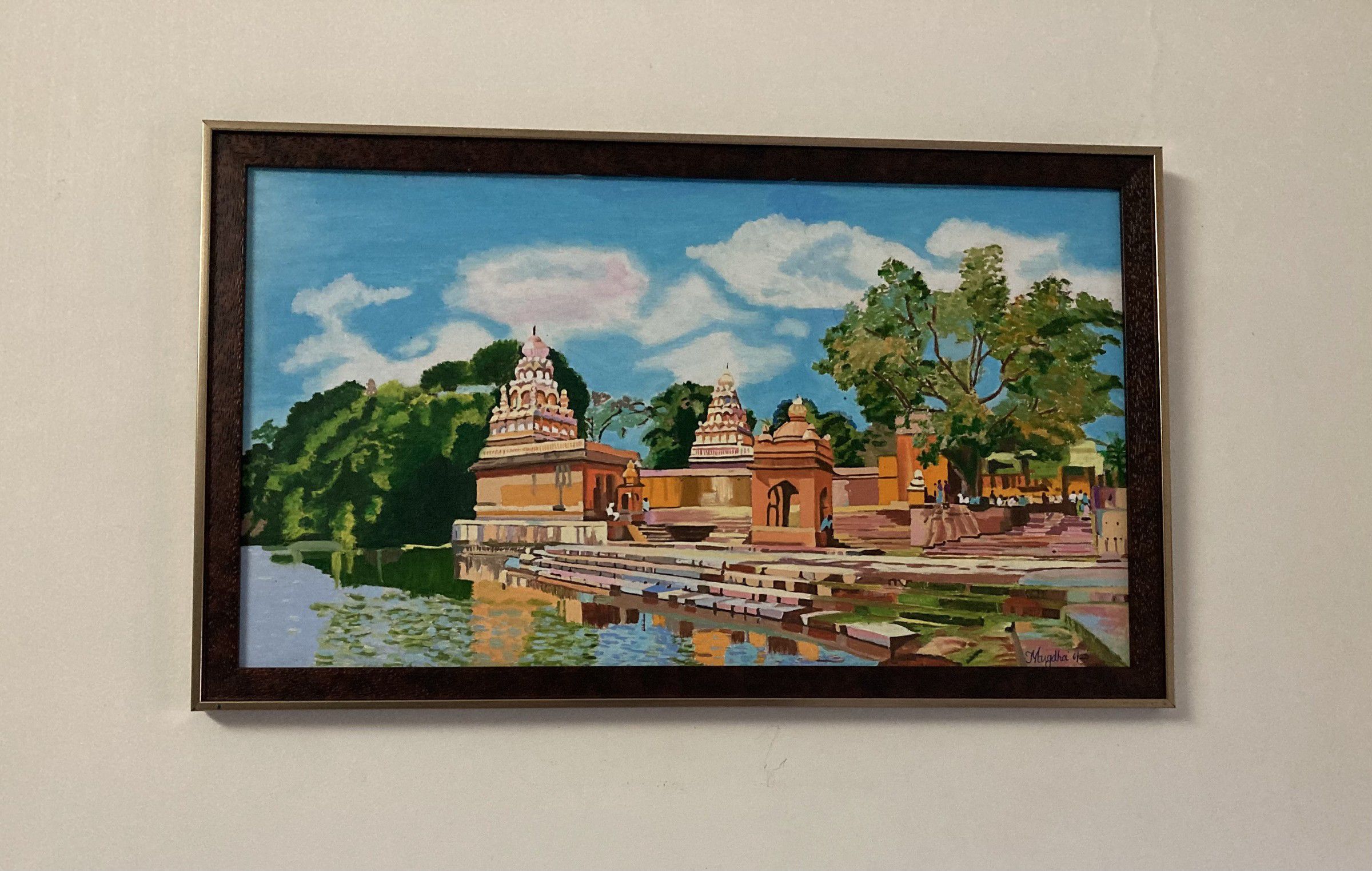 Framed painting of temples on a riverbank, with stone steps down to the water and trees under a blue sky