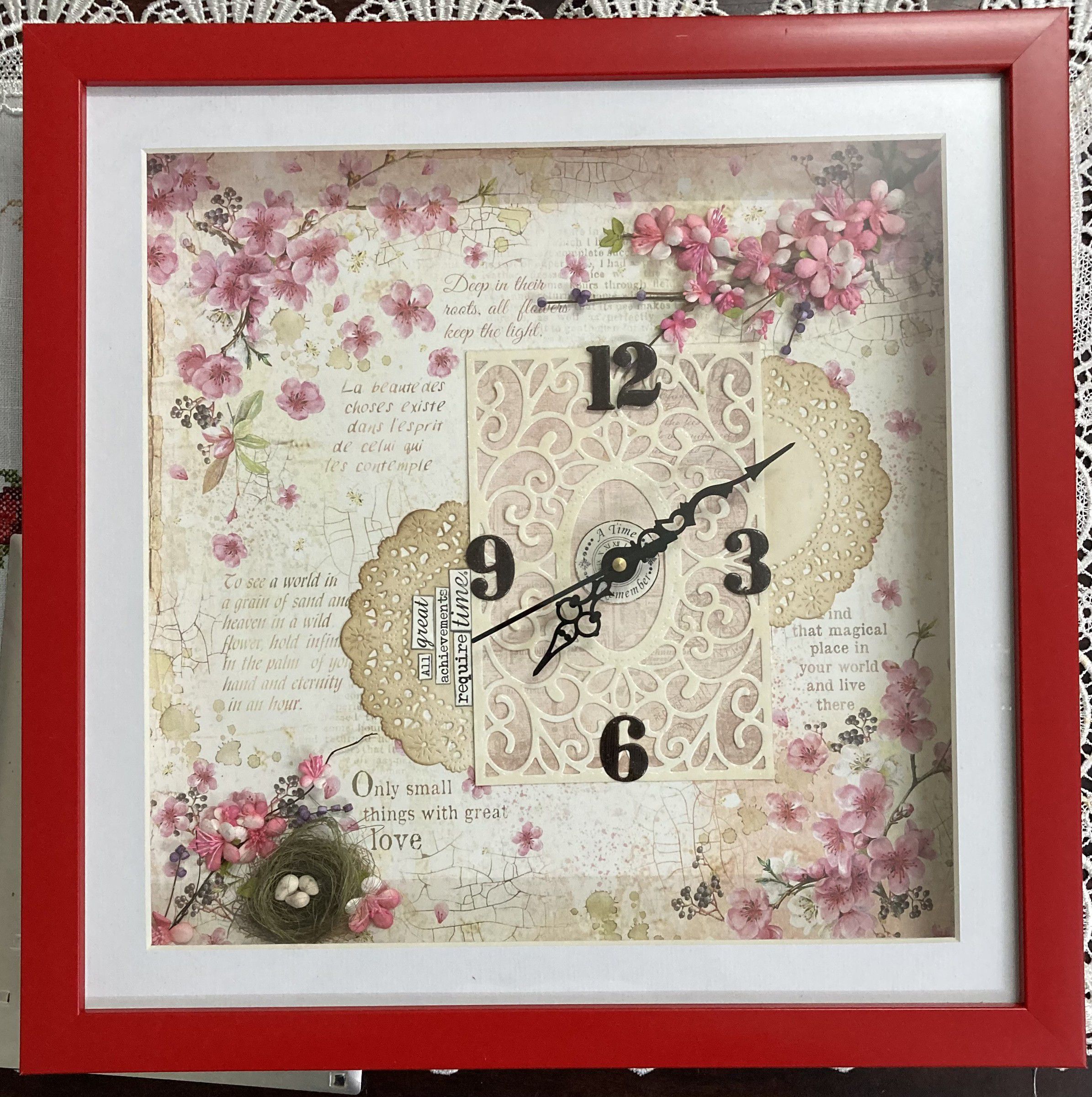 Red shadow-box frame holding a clock on a cherry-blossom background, with a lace doily and a tiny nest of eggs