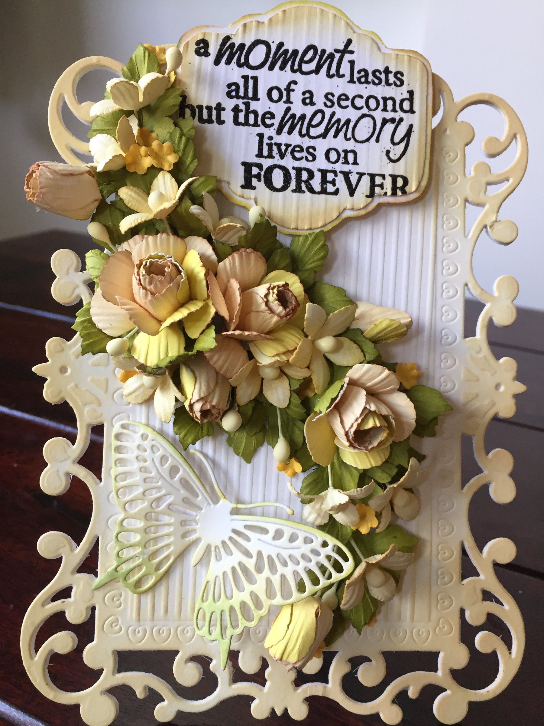 Card with cream and yellow paper roses, a butterfly and a tag reading a moment lasts all of a second but the memory lives on forever
