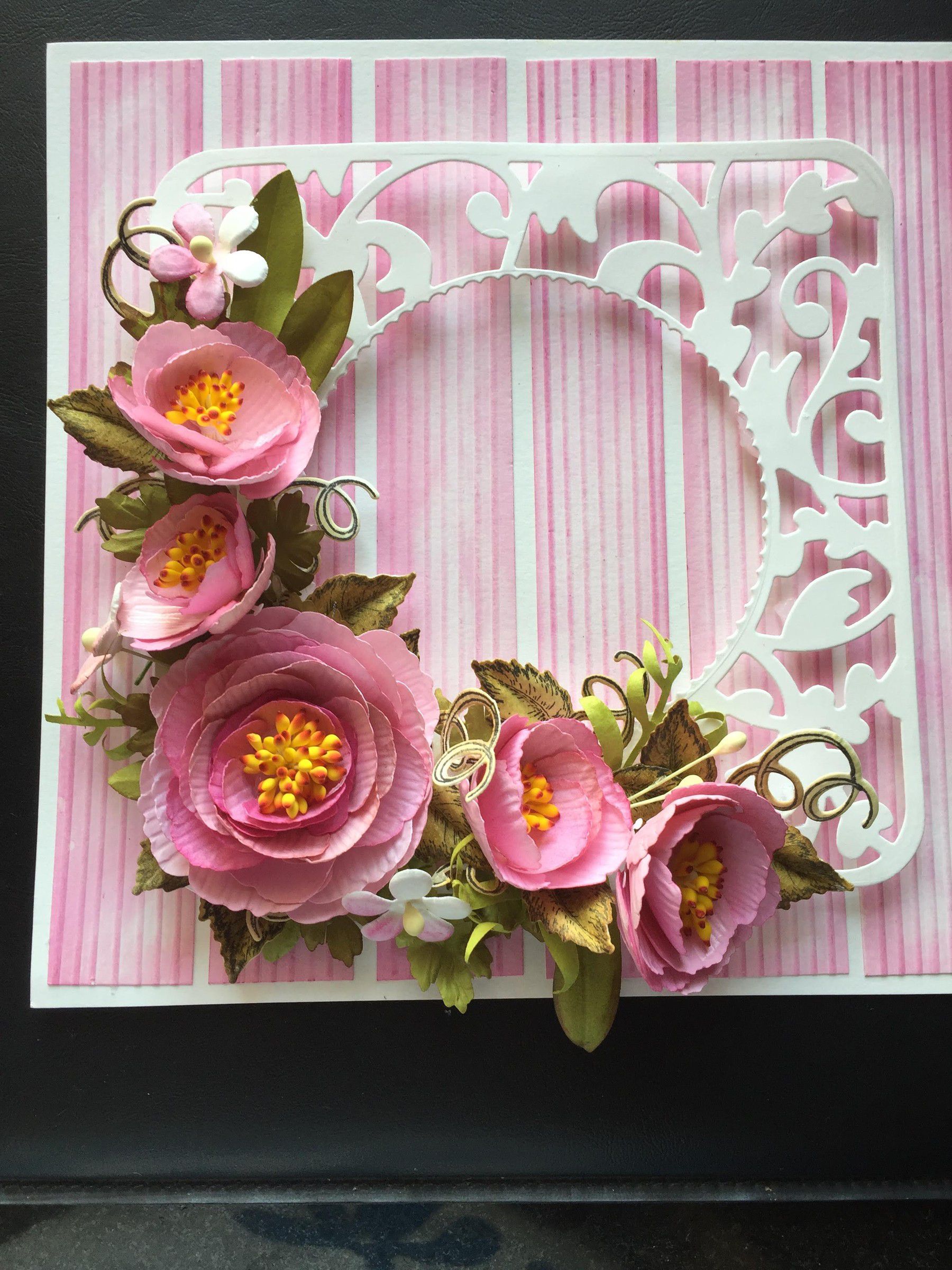 Card with pink paper peonies and green leaves over a white filigree frame on pink stripes