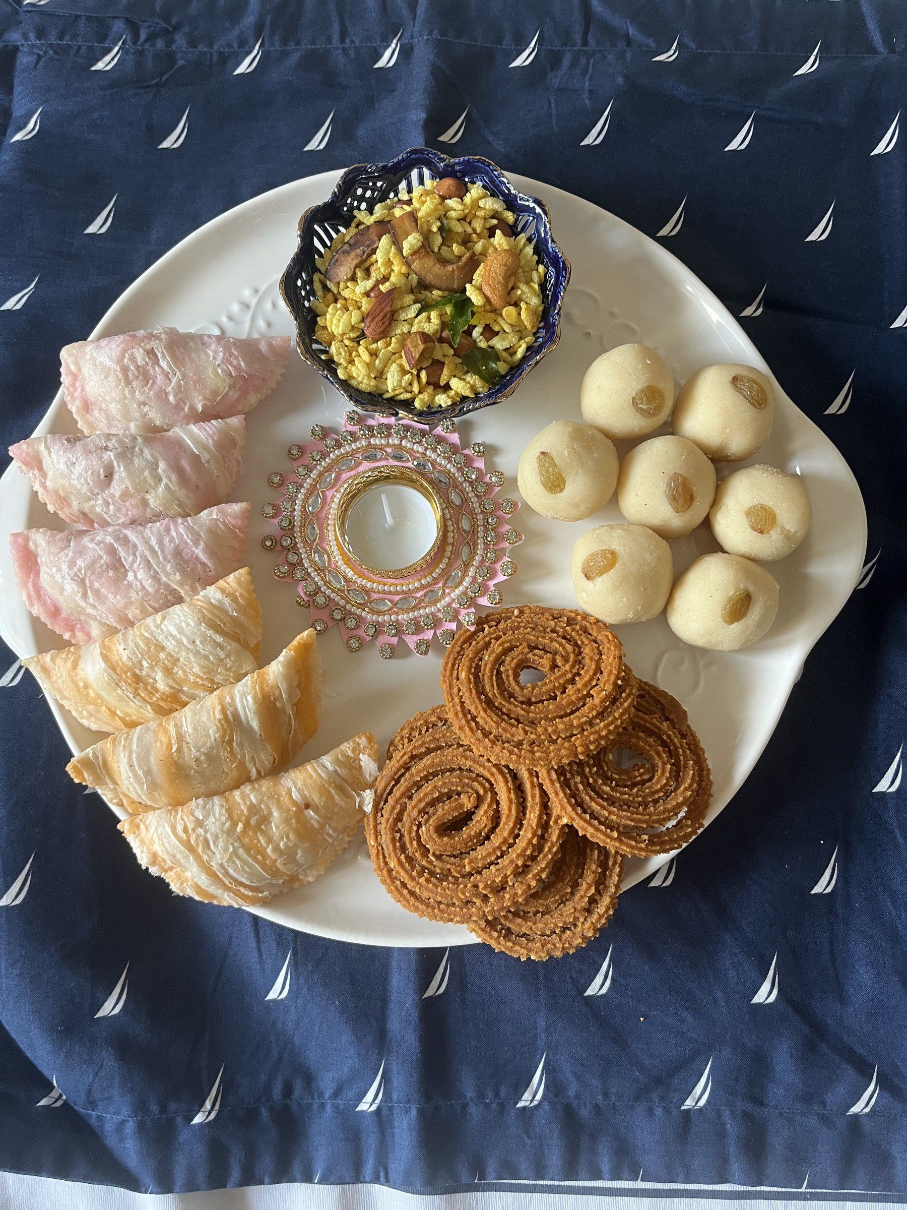Festive platter of pink and plain karanji, chakli, ladoos and a bowl of chivda around a decorative diya