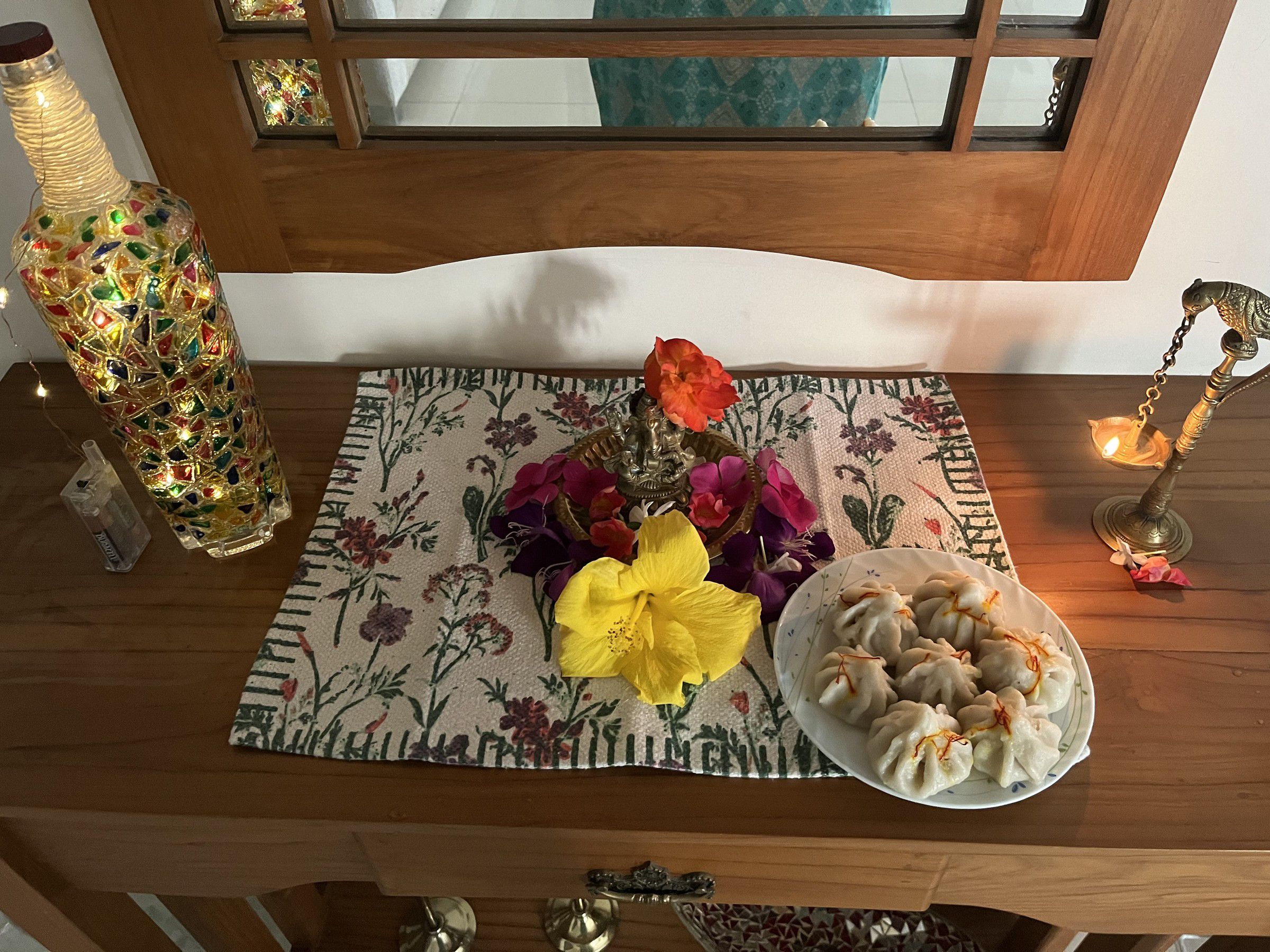 Plate of steamed modak beside a small Ganpati idol, flowers and a brass lamp on a wooden table