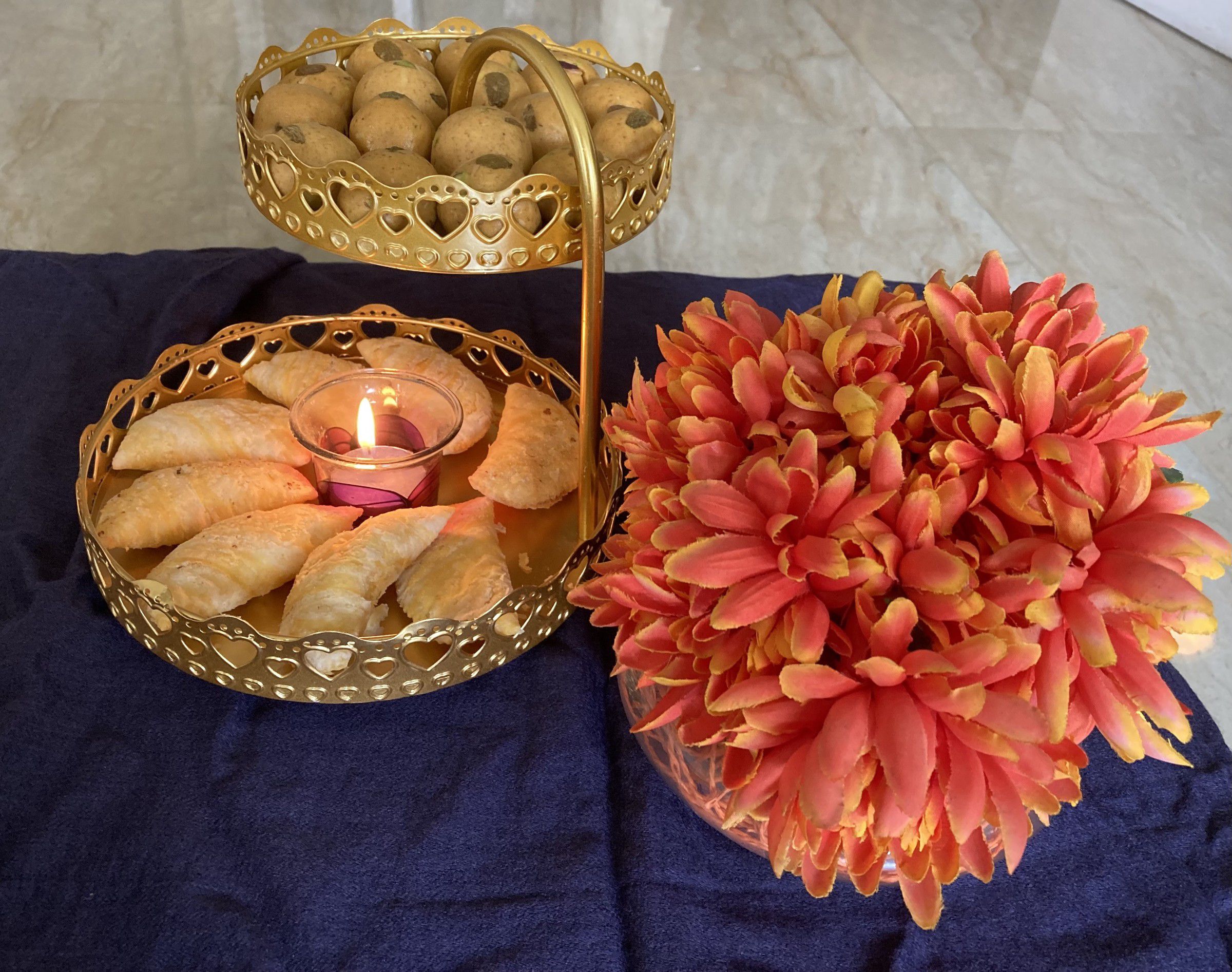 Two-tier gold stand holding ladoos and karanji with a lit tealight, beside orange chrysanthemums