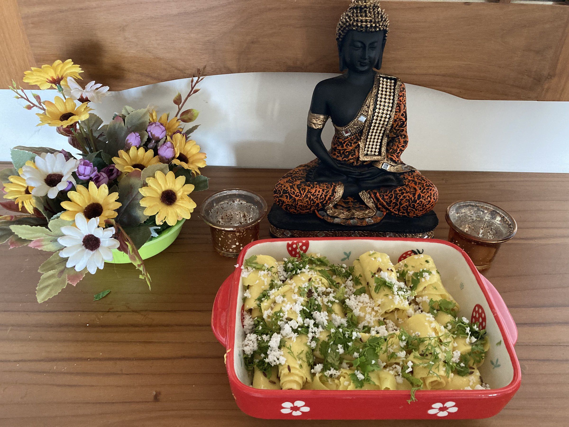 Red dish of khandvi rolls garnished with grated coconut and coriander, beside a Buddha figure and flowers