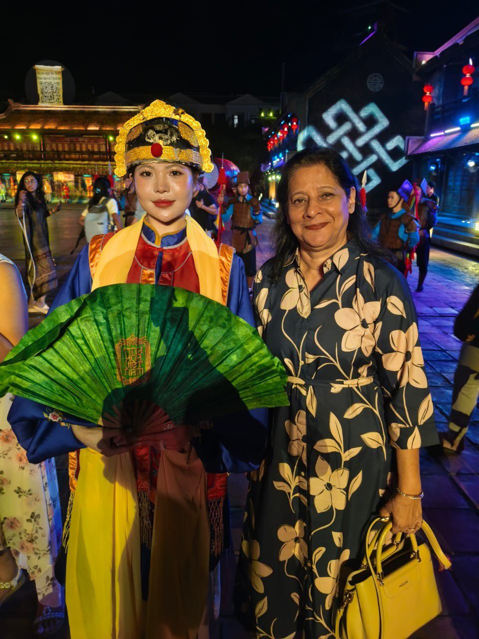 Mugdha at night beside a performer in an ornate costume holding a green fan, on a lantern-lit street