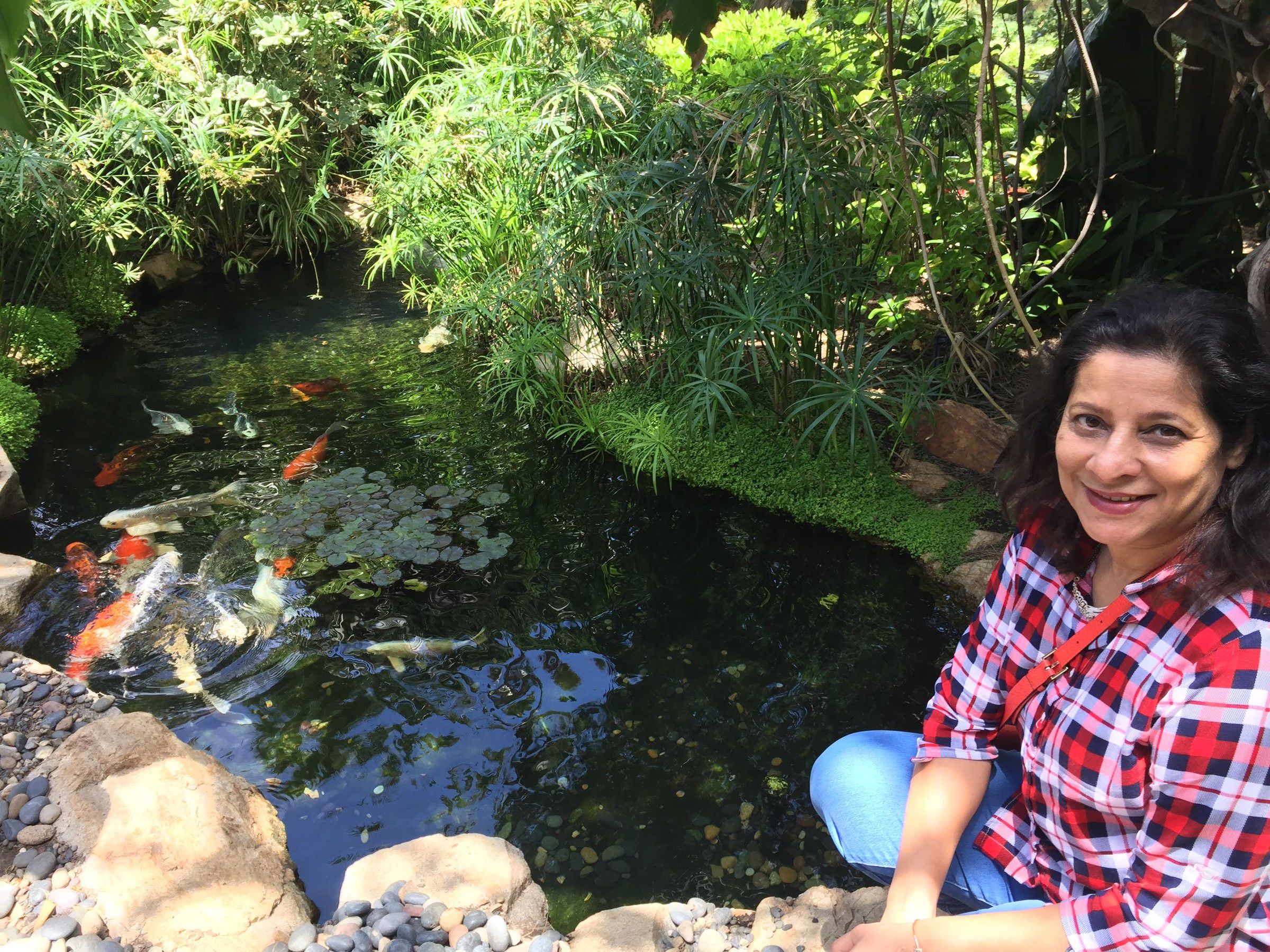 Mugdha smiling beside a koi pond surrounded by lush plants